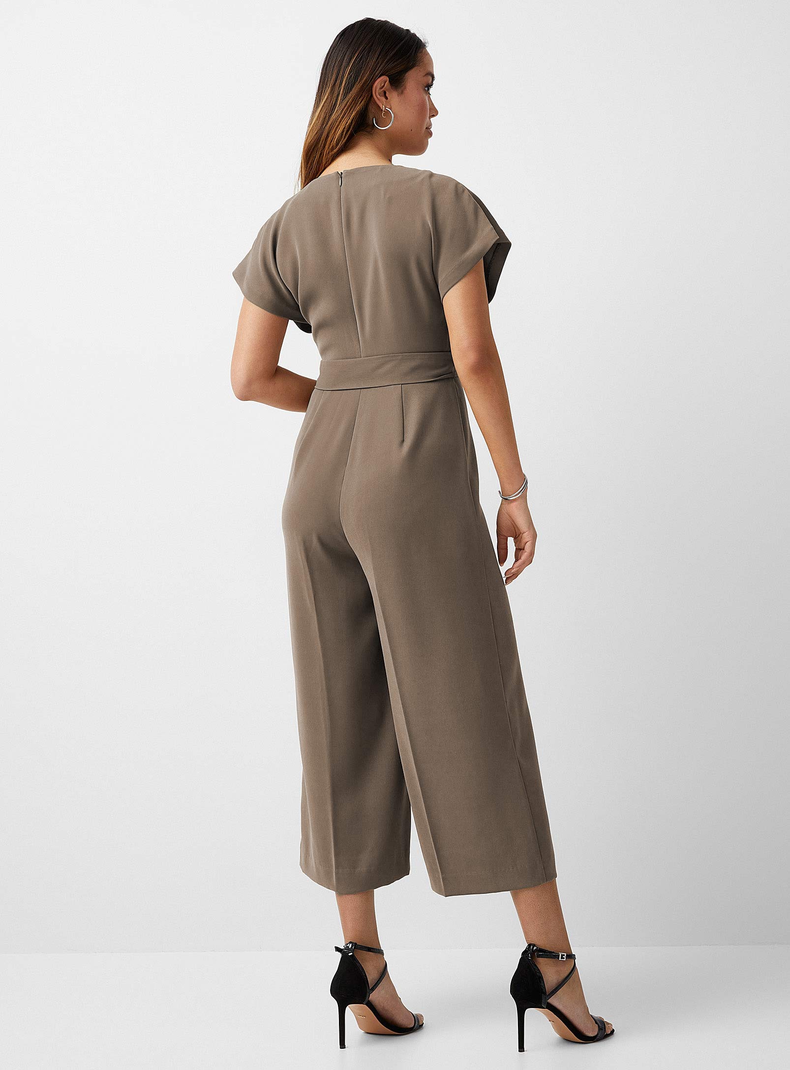 Fashion-forward woman showcasing a taupe jumpsuit with a flattering V-neckline and short sleeves. The outfit is cinched at the waist with a tie belt, leading into wide-legged cropped pants, creating a sleek silhouette